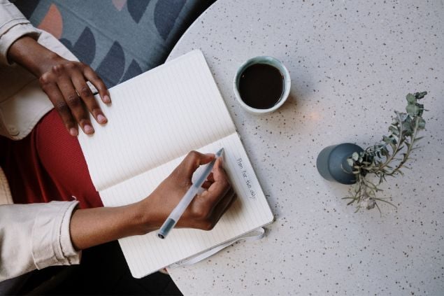 Imagem de uma pessoa escrevendo em sua agenda na mesa, e ao lado uma xícara de café