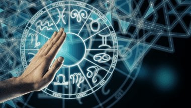 Há uma Roda do Zodíaco com os símbolos dos signos em volta. Na frente, há a mão de uma pessoa.