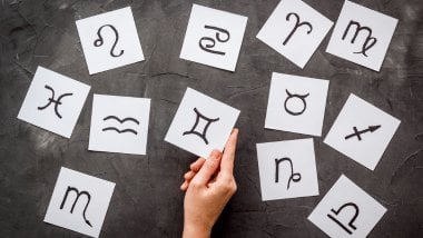 Mão segurando um cartão com símbolo de Gêmeos, cercada por outros cartões com signos do zodíaco sobre fundo cinza.