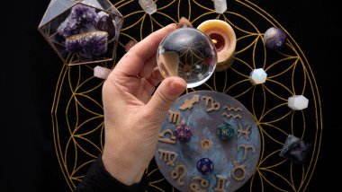 Mão segurando uma esfera transparente com símbolos astrológicos, cercada por cristais e elementos místicos sobre fundo escuro.