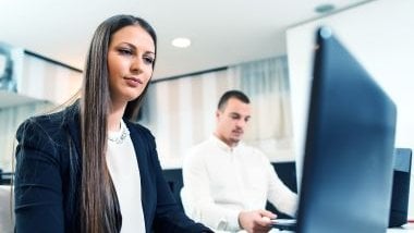 Imagem de um homem e uma mulher em uma sala de um escritório. Ambos estão em suas ilhas de trabalho olhando para os computadores.