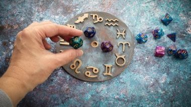 Imagem de uma mão feminina manipulando pedras de cristais coloridas sobre uma mandala astrológica com os elementos dos 12 signos.