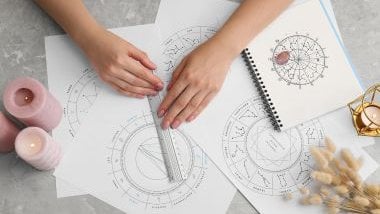 Imagem das mãos de uma astróloga elaborando um mapa natal. Na mesa, várias folhas, velas acesas na cor rosa.