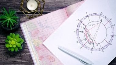 Imagem de uma mandala astrológica sendo elaborada. O papel onde ela está sendo desenhada fica sobre um caderno de folhas rosas disposto em uma mesa de madeira.