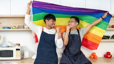 Imagem de um casal LGBTQIAPN+ segurando a bandeira da causa. Eles estão em uma cozinha dentro de casa.