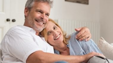 Imagem de um casal maduro, abraçados e sentados em uma poltrona na sala.