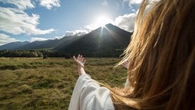 Imagem de uma mulher de costas olhando para um monte verde em frente ao Sol.
