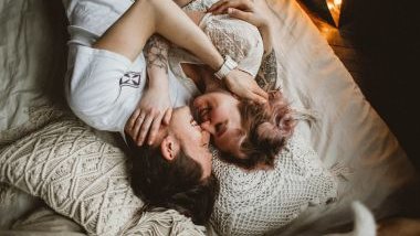 Imagem de um casal deitado sobre uma cama. Eles estão abraçados, conversando e ao mesmo tempo, sorrindo.