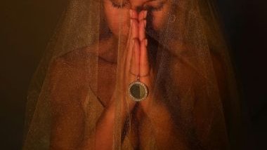 Imagem do rosto de uma mulher coberto com um véu dourado com as mãos unidas em sinal de oração, segurando uma corrente prata com pedras.