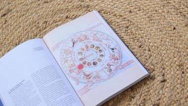 Imagem de um livro aberto sobre uma toalha feita de palha. Na página, uma imagem com uma mandala astrológica e os 12 signos do zodíaco.
