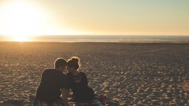 Imagem de um casal, homem e mulher, sentados na praia, em frente ao pôr do Sol.