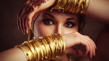 Imagem do rosto de uma mulher negra usando roupas em tons de dourado e com colares de contas douradas, flores discretas e adornos, simbolizando Oxum Ijimu.