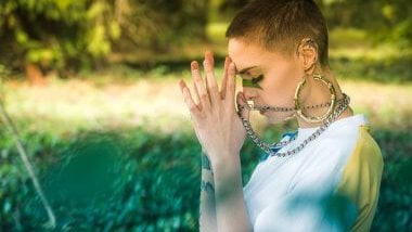 Imagem de uma mulher de cabelos bem curtinhos, usando brincos de argola e corrente grossa, orando em um campo aberto, todo gramado, simbolizando a Iyalorixá, guia espiritual feminina do Candomblé.