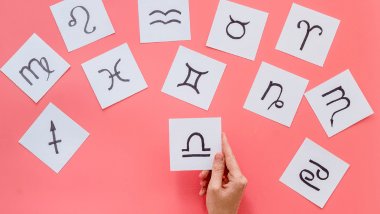 Cada símbolo de um signo do zodíaco está desenhado em um pedaço de papel. Os papéis estão espalhados em cima de um fundo cor de rosa.