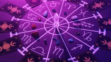 No foco da imagem, há uma Roda do Zodíaco e, em cima dela, há pedras e cartas de Tarot.