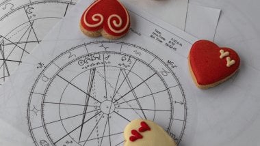 No foco da imagem, há uma Roda do Zodíaco desenhada em um papel com os símbolos dos signos e há biscoitos em formato de coração ao redor.