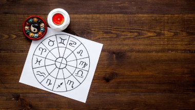 Há uma roda do zodíaco com os símbolos dos signos desenhada em papel sulfite em cima de uma mesa. Ao redor, há velas.