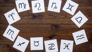 Cada símbolo de um signo do zodíaco está desenhado em um pedaço de papel. Os papéis estão espalhados em cima de uma mesa de madeira.