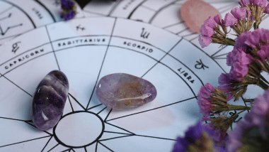 Há três rodas do zodíaco com os símbolos dos signos desenhadas em papel sulfite em cima de uma mesa. Ao redor, há pedras e flores.