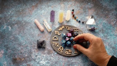 No foco da imagem, há uma Roda do Zodíaco com os símbolos dos signos e há dados, pedras e cristais ao redor.