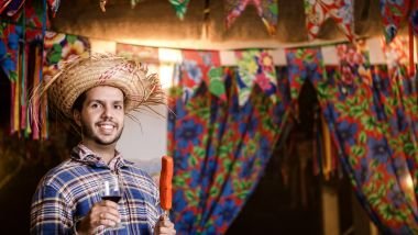 Imagem de um homem vestido de caipira, segurando um espeto de salsicha assada e uma taça de vinho, curtindo uma festa junina.