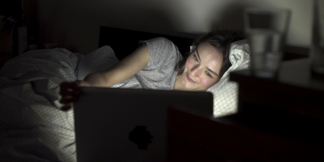 Mulher deitada em cama sorri ao observar a tela do notebook que também está sobre a cama.