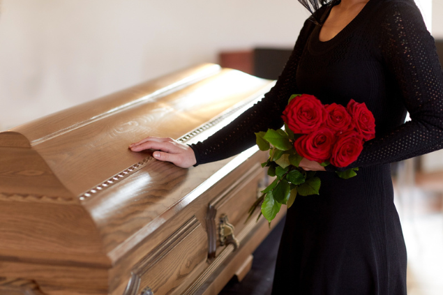 Mulher ao lado de um caixão fechado, segurando rosas vermelhas.