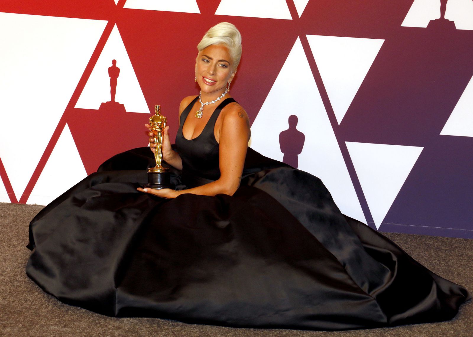 Lady Gaga sentada no chão usando vestido segurando seu prêmio do Oscar