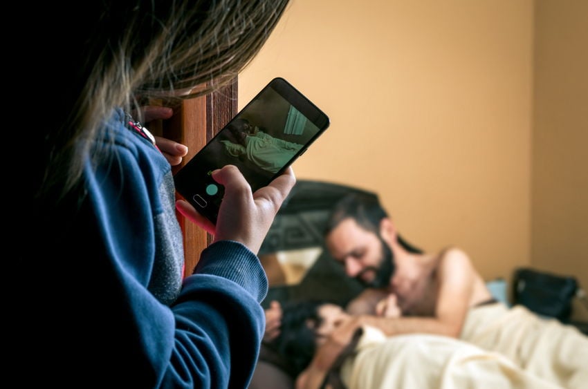 Mulher branca com celular na mão fotografando casal deitado numa cama.
