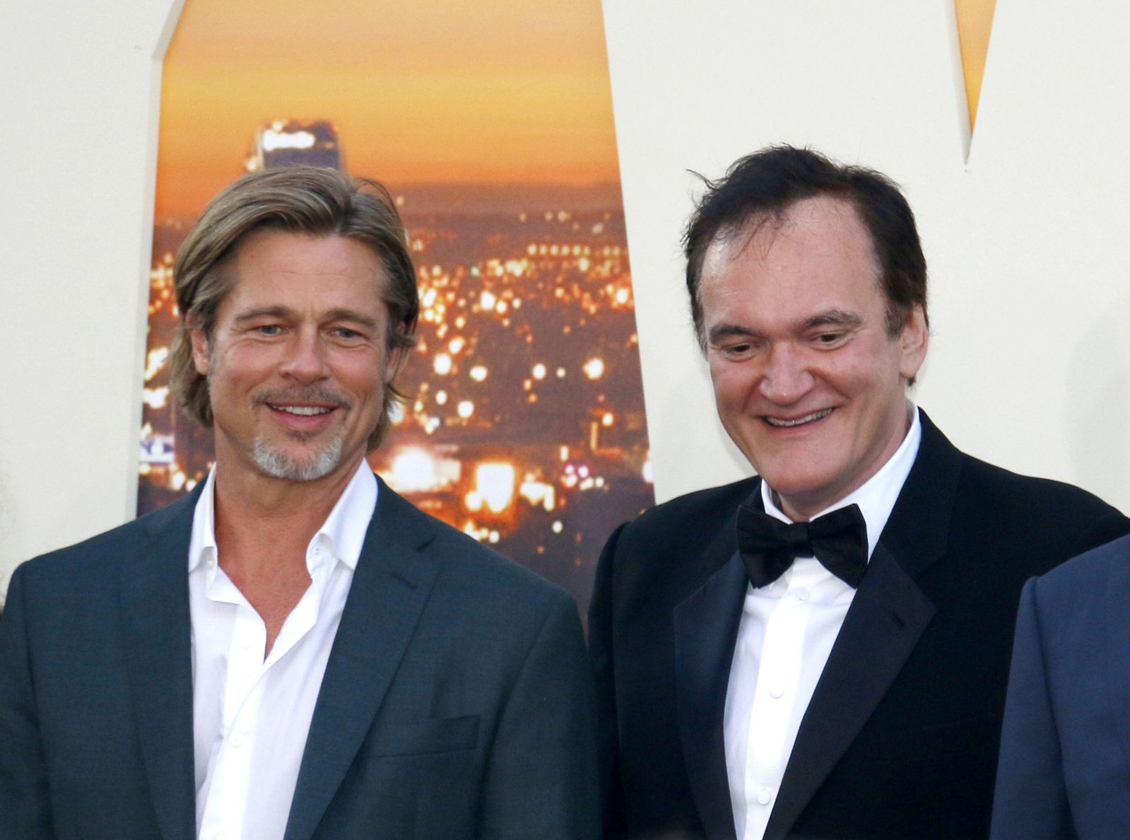 Quentin Tarantino sorrindo e usando terno ao lado de um ator 