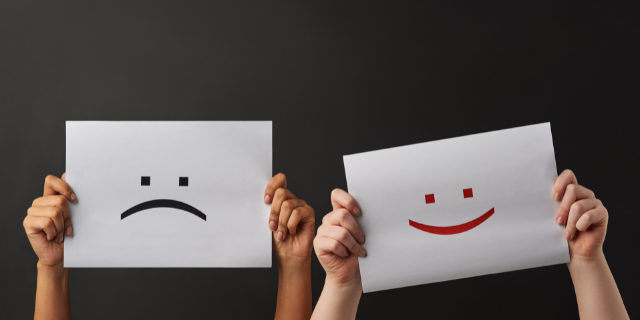 Duas pessoas segurando um papel com emoji. À esquerda, uma com um rosto triste desenhado e à direita, uma com um rosto feliz desenhado. 