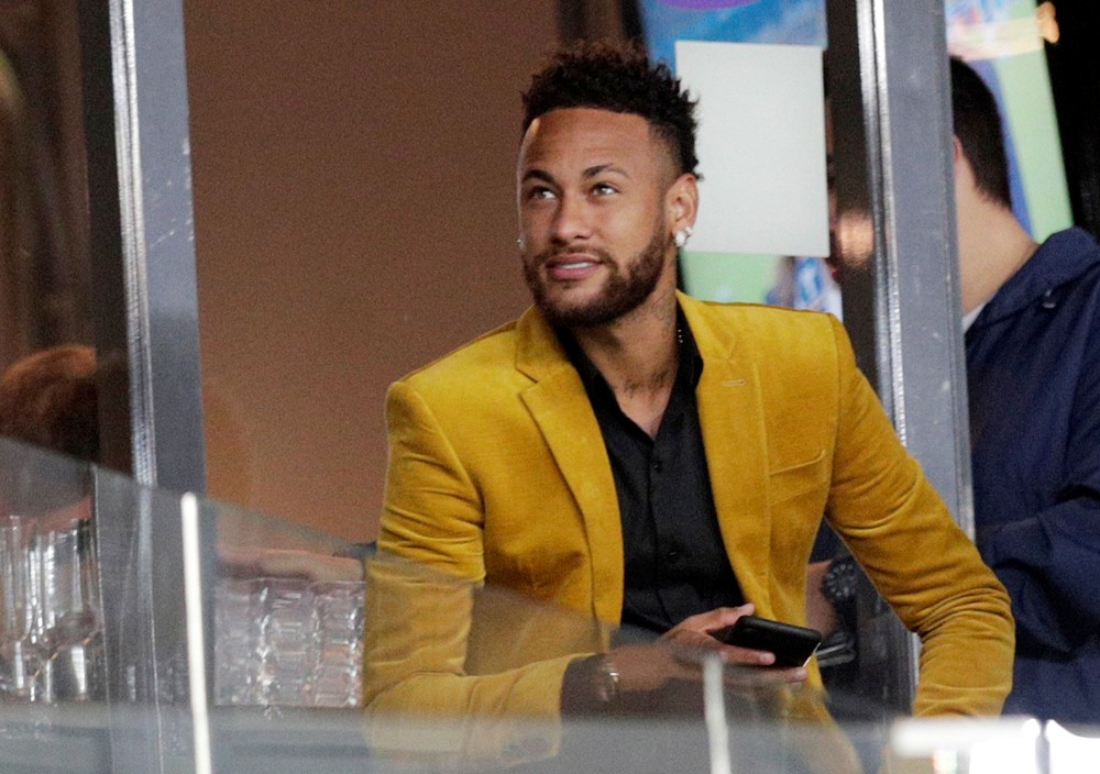 Neymar em estádio, assistindo jogo, vestindo um terno amarelo e usando o celular.