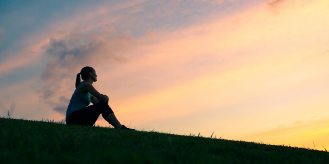 Silhueta de mulher sentada na grama observando o por do sol