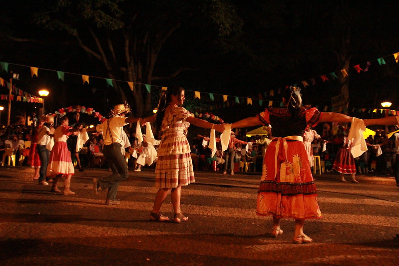 Pessoas dançando quadrilha, de mãos dadas, durante a noite.