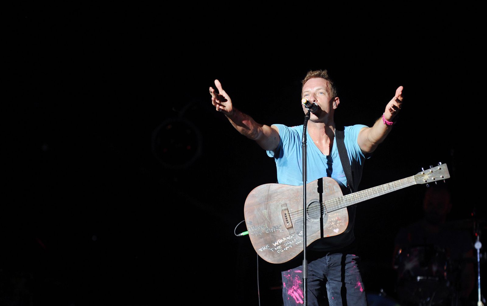 Chris Martin cantando no palco com violão e de braços abertos