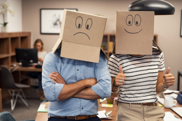 Duas pessoas lado a lado com papelões na cabeça. À esquerda, uma com um rosto triste desenhado e à direita, uma com um rosto feliz desenhado. 