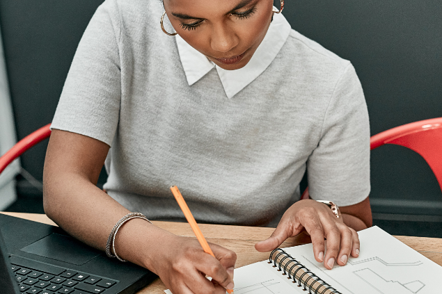 Mulher escrevendo em um caderno enquanto trabalha em frente a um notebook