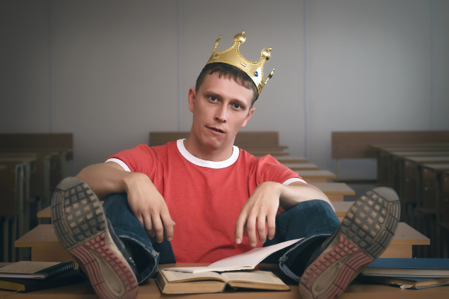 Homem sentado com as duas pernas em cima de uma carteira e com uma coroa e rei na cabeça
