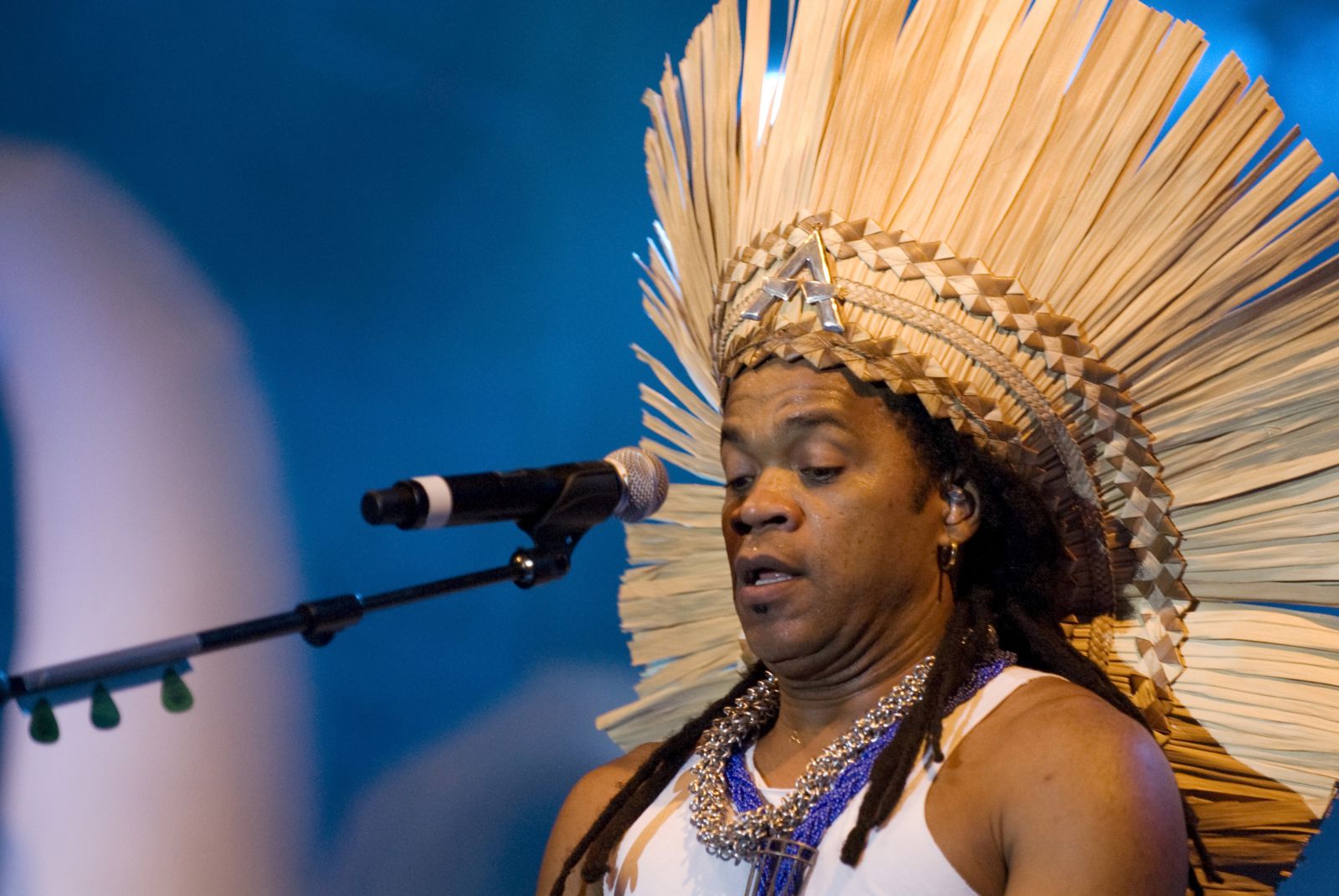 Carlinhos Brown tocando e cantando no palco usando cocar na cabeça