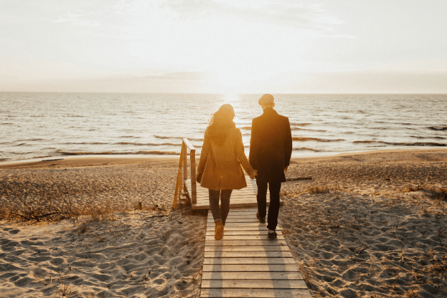 Casal na praia andando de mãos dadas em direção ao pôr-do-sol