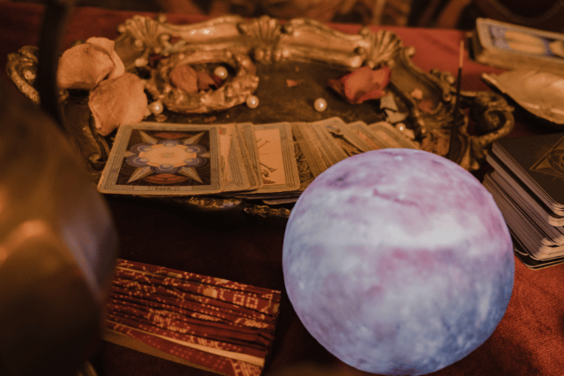 Faruk Tokluoglu @faruk-tokluolu / Studio Turkey / Canva Há várias cartas de Tarot viradas para baixo em cima de um prato sobre uma mesa. Ao lado, há uma bola de cristal e outros objetos.