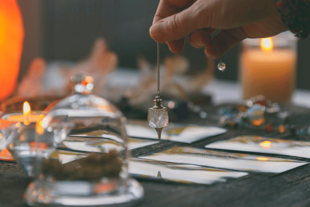 Humonia / Getty Images / Canva Algumas cartas de Tarot viradas para baixo estão em destaque na imagem. Uma taróloga segura um cristal em cima das cartas. Ao lado, há velas e outros objetos.
