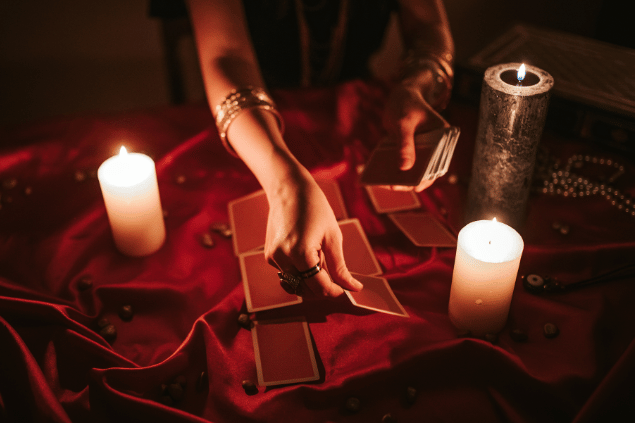 As mãos de uma taróloga segurando e posicionando cartas de Tarot viradas para baixo estão em destaque na imagem. Há velas ao lado.