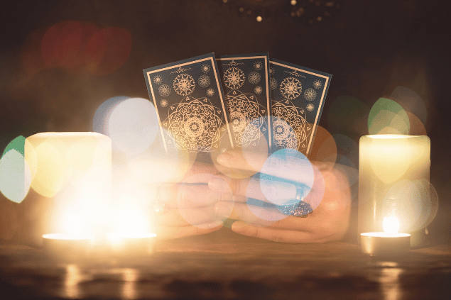 HappyNati / Getty Images / Canva As mãos de uma taróloga segurando cartas de Tarot estão em destaque na imagem. Ao lado, há velas.