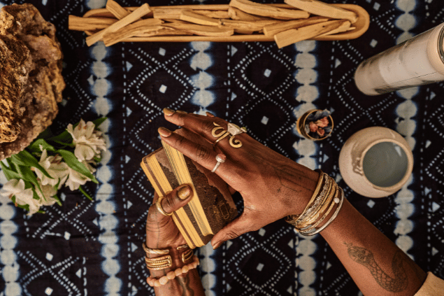 B Dukes / B Dukes / Canva As mãos de uma taróloga segurando cartas de Tarot viradas para baixo estão em destaque na imagem. Ao lado, há uma vela e outros objetos.