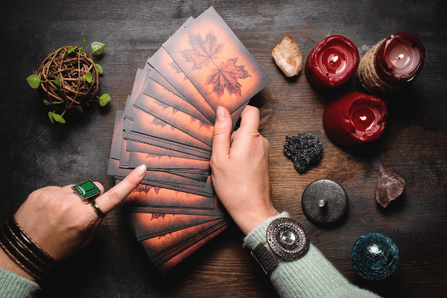 As mãos de uma taróloga segurando cartas de Tarot viradas para baixo estão em destaque na imagem. Ao lado, há velas e outros objetos.