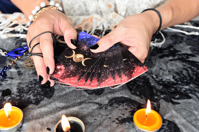 Bigc Studio / Canva As mãos de uma taróloga segurando cartas de Tarot viradas para baixo estão em destaque na imagem. Ao lado, há velas e outros objetos.