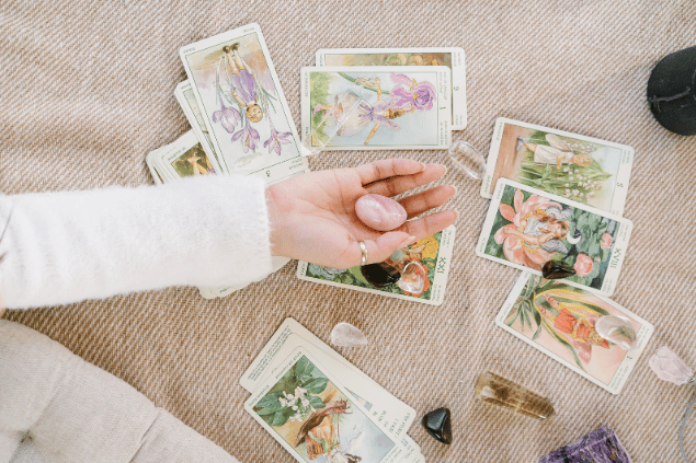 Mikhail Nilov / Pexels / Canva A mão de uma taróloga segurando uma pedra está em destaque na imagem. Embaixo, há algumas cartas de Tarot espalhadas.