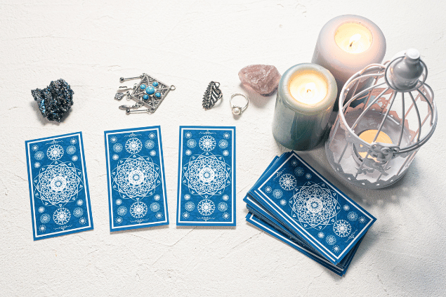 Algumas cartas de Tarot viradas para baixo estão em destaque na imagem. Ao lado, há três velas e outros objetos.