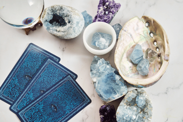 Vetrestudio / Canva Algumas cartas de Tarot viradas para baixo estão em destaque na imagem. Ao lado, há cristais e outros objetos.